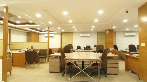 Interior of a contemporary bank, showcasing an open floor plan with multiple workstations equipped with computers, ergonomic chairs, and wooden partitions. A central meeting table surrounded by chairs is featured, and the space is illuminated by overhead ceiling lights. The design employs a neutral color palette, contributing to a professional and modern atmosphere